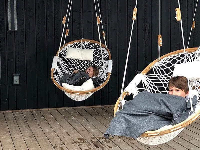 Two young boys covered with blankets sitting in Cobble Mountain Summit Hammock chairs. with white pillows. The boys are laughing