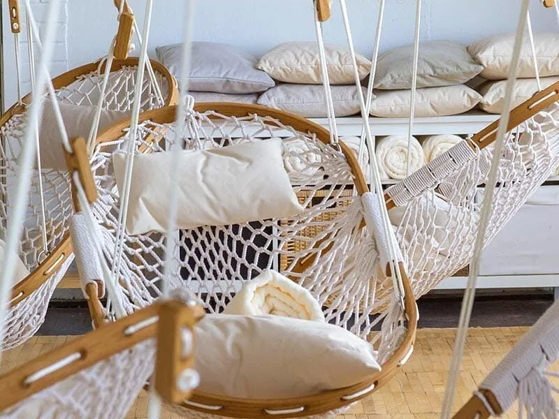 A close up of Cobble Mountain Original Hammock Chairs with white pillows hanging in a loft. in the back ground are white pillows and blankets stored on a bench.